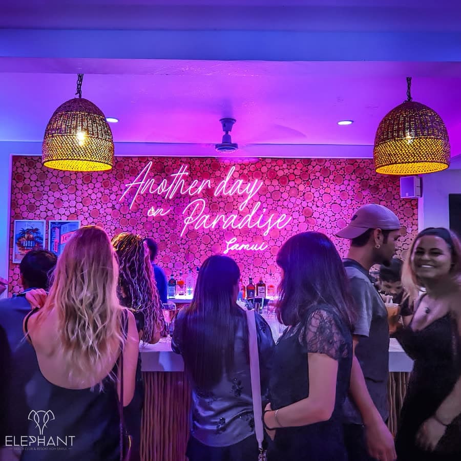Guests at the Elephant Bar counter under the pink neon sign reading Another day in Paradise, rattan pendants overhead, Koh Samui