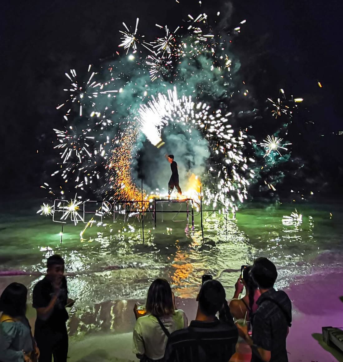 Fire performer on a frame above the water throwing a wide burst of sparks over the bay while guests watch from the sand at Elephant Beach Club, Chaweng Beach