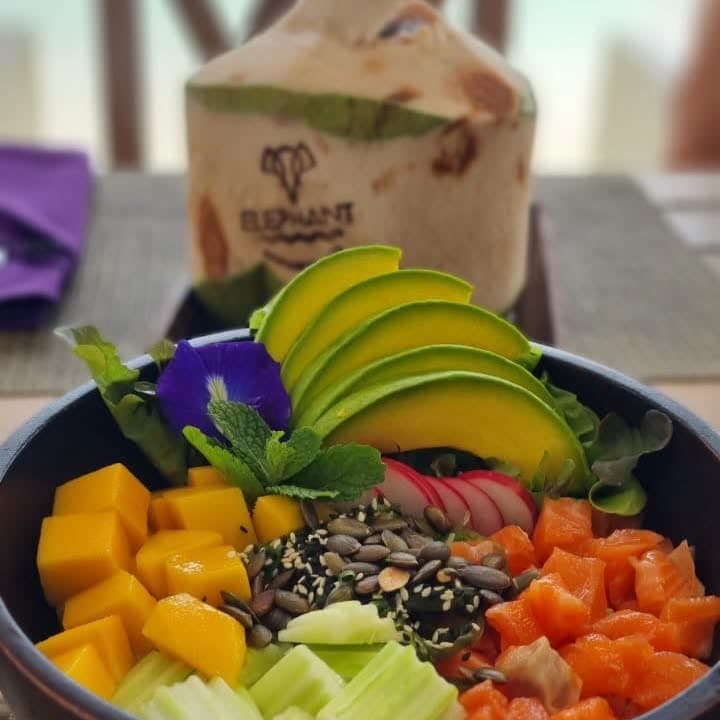 Salmon poke bowl with mango, avocado, cucumber and sesame seeds, an Elephant-branded coconut behind it, Chaweng Beach