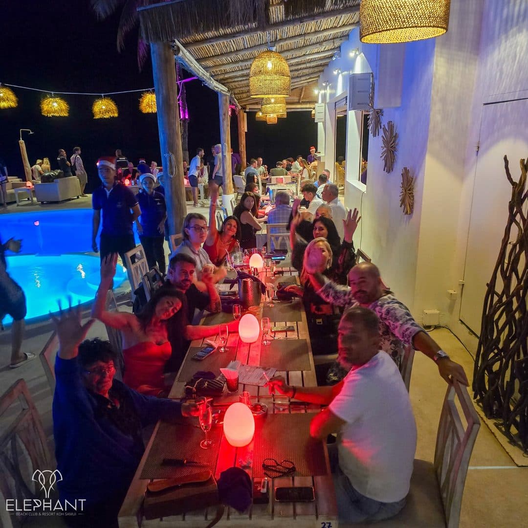 Guests celebrating around a long table by the pool at Elephant's beachfront restaurant at night, red mood lighting, staff and diners raising a toast, Chaweng Beach, Koh Samui