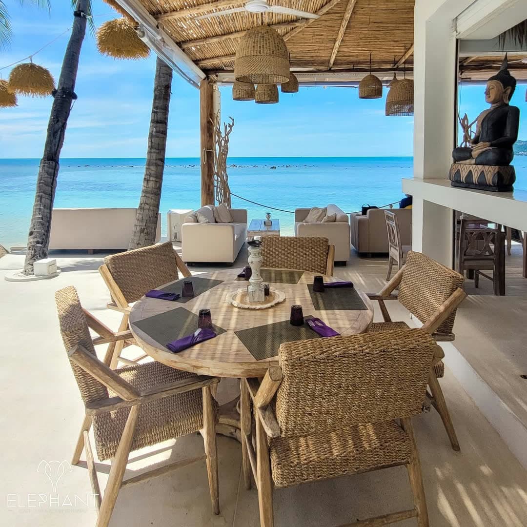 Round driftwood table set with purple napkins and rattan chairs, a seated Buddha and the open sea behind, at Elephant beachfront restaurant