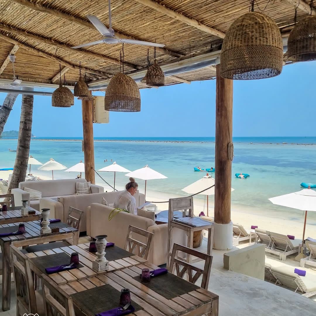 The covered terrace at lunchtime, wooden tables under rattan pendants, looking out to the sunbeds and the turquoise water of Chaweng Bay