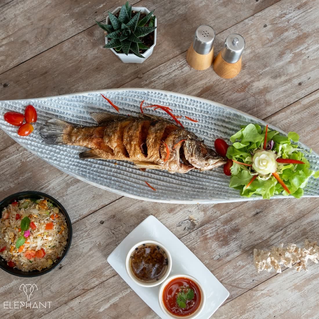 Whole fried sea bass on a long ceramic platter with salad, fried rice and two dipping sauces, seen from above at Elephant beachfront restaurant